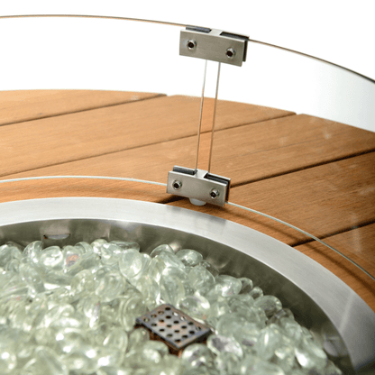 This close-up of the gas fire pit focuses on the metal clamps securing the transparent glass wind guard and the surrounding tabletop material. The metal clamp consists of two rectangular parts, fastened with two screws to the circular glass wind guard. The bottom of the clamp is attached with another screw to the light brown faux wood slat tabletop. The tabletop is made of parallel wood slats with subtle gaps between them, creating a natural wood grain effect.