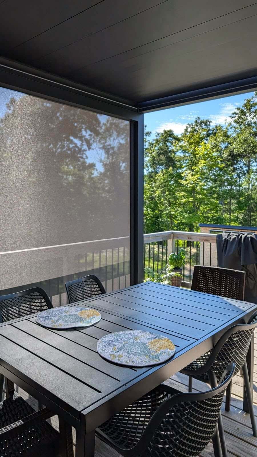 The BON Pergola features a fully closed louvered top with a sturdy deep gray frame of thick posts and beams. Light gray motorized blinds are installed on the sides, some partially lowered to provide shade and privacy. The fabric appears semi-translucent, effectively blocking direct sunlight while allowing filtered light through. Through the partially lowered blinds, you can catch glimpses of the outside view—lush green trees and a clear blue sky.