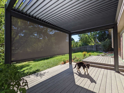 A black Bon Pergola, 13 ft by 19 ft, with motorized louvered roof panels, stands over a patio decked in light gray wood. One side of the pergola features a dark-colored sunshade screen. The patio is open and uncluttered, with just a small dog wandering through the sunlight.