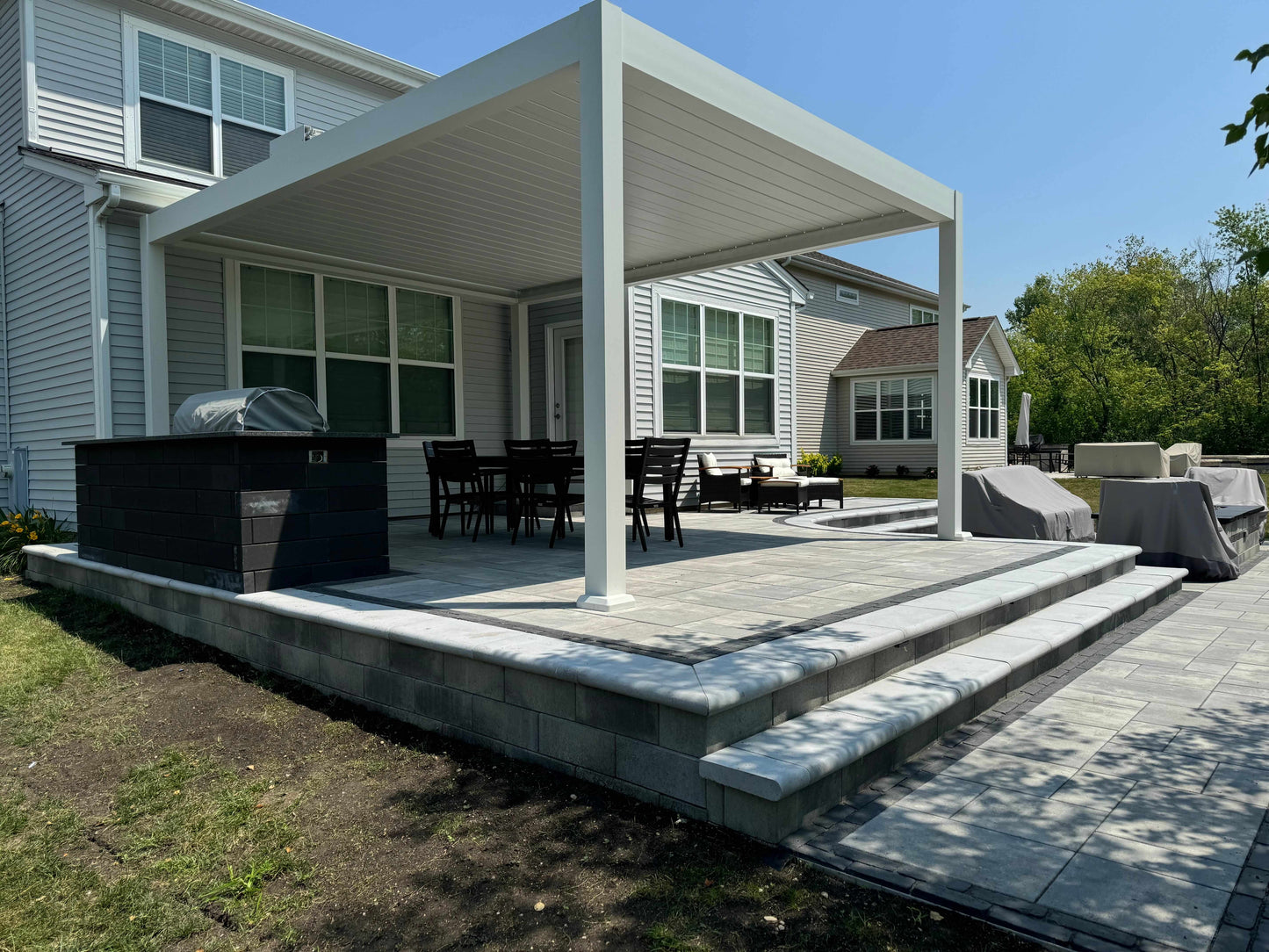A white BON PERGOLA is installed on the residential backyard terrace, with the louvered top fully closed, forming a solid white roof. The pergola's structure consists of sturdy white columns and beams, giving it a sleek, modern look. One side of the pergola is attached to the home's exterior wall. The terrace is furnished with outdoor furniture, including a black rectangular dining table and matching chairs, providing shade and creating a pleasant outdoor space for activities.