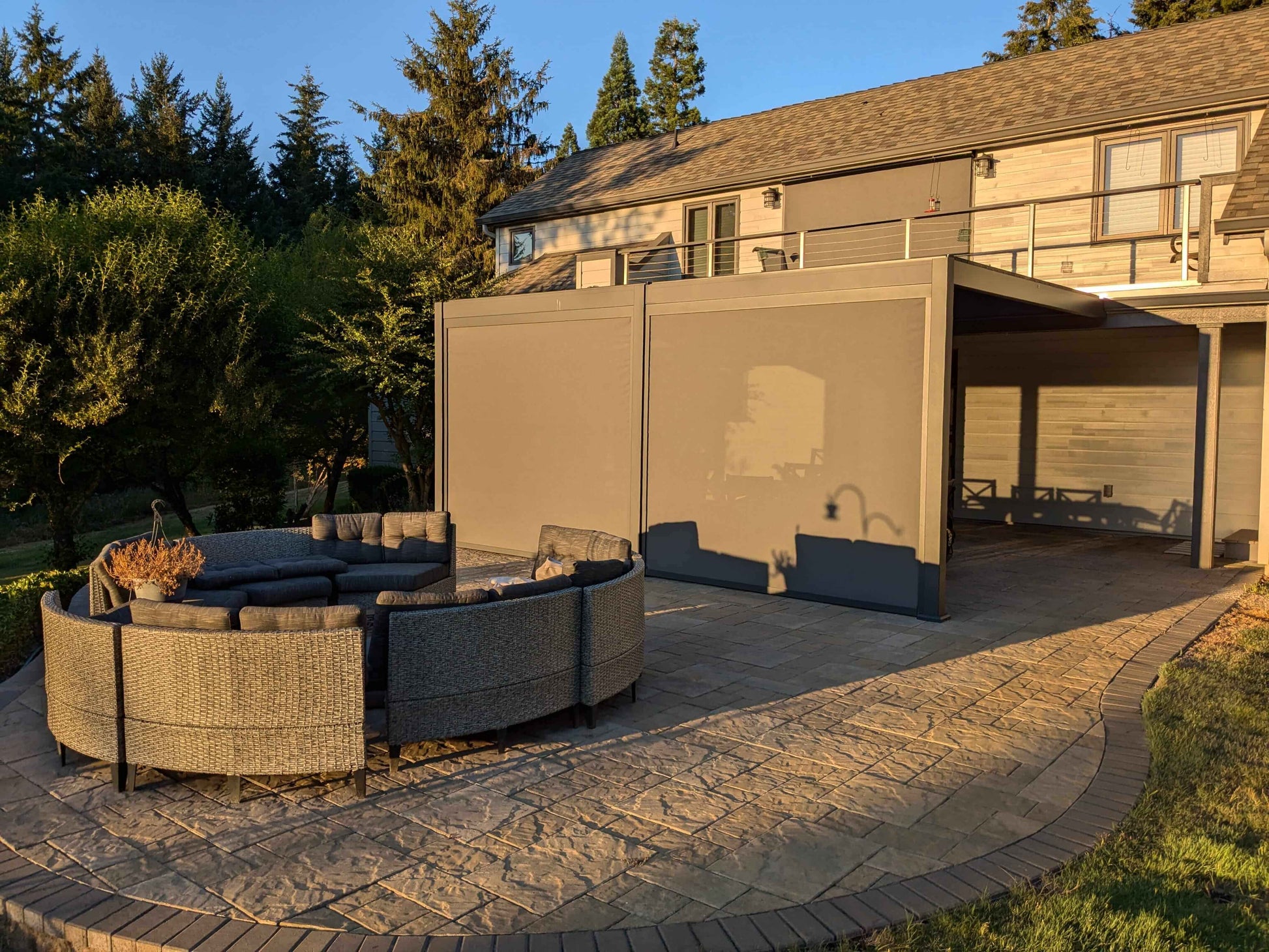 As the sun sets, the Villa Pergola’s deep gray louvers are fully closed, forming a sleek roof. The sturdy frame is matched with light gray motorized blinds, partially lowered for shade and privacy. The fabric is semi-translucent, blocking direct sun while letting in soft light. Beneath, a curved gray outdoor sofa with dark gray cushions offers a cozy lounge spot. The patio sits beside a light wood-paneled house exterior with windows and a balcony, surrounded by lush lawn and trees.