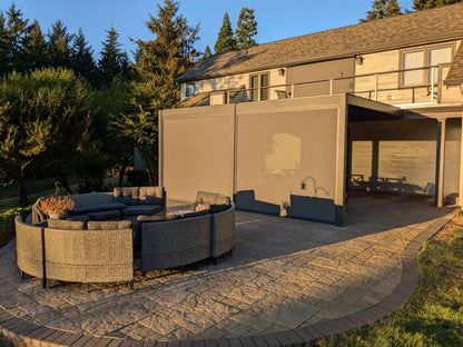 As the sun sets, the Villa Pergola’s deep gray louvers are fully closed, forming a sleek roof. The sturdy frame is matched with light gray motorized blinds, partially lowered for shade and privacy. The fabric is semi-translucent, blocking direct sun while letting in soft light. Beneath, a curved gray outdoor sofa with dark gray cushions offers a cozy lounge spot. The patio sits beside a light wood-paneled house exterior with windows and a balcony, surrounded by lush lawn and trees.