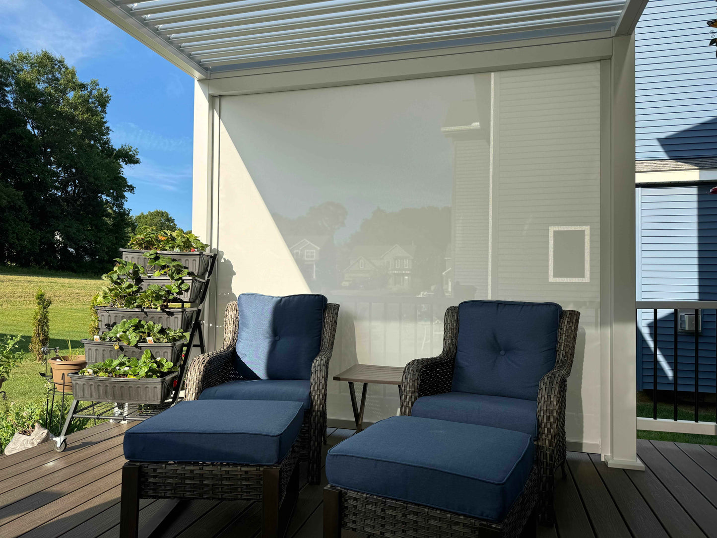 The terrace features white motorized sunshade blinds, partially lowered for shade. In the center, two outdoor rocking chairs with deep blue cushions and backrests are framed in dark brown woven material. Each chair has a matching footstool with the same woven design and blue cushion. Between the chairs is a small light brown side table. This cozy, well-arranged outdoor space offers shade and comfort, making it an ideal spot for relaxation.