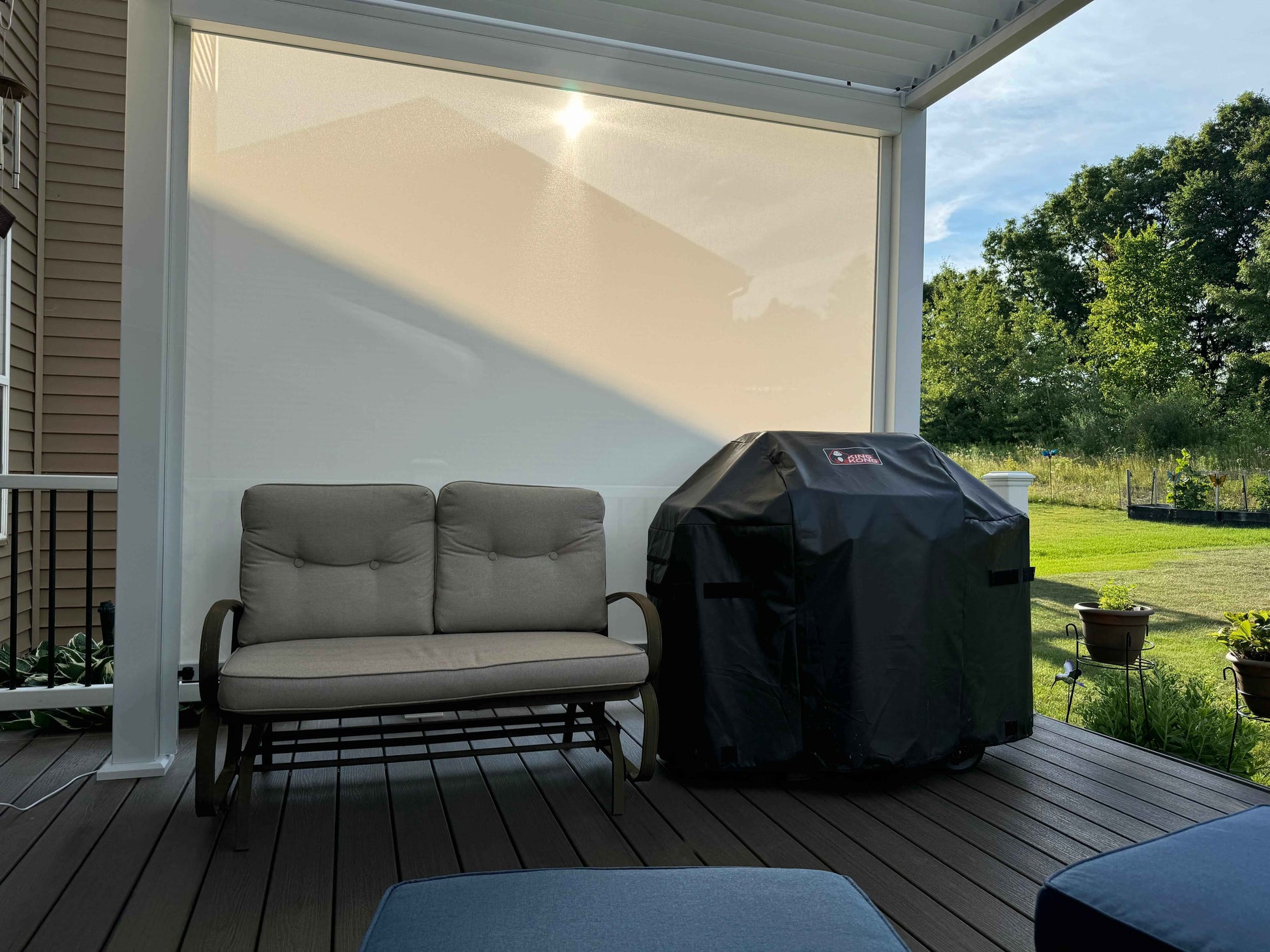 The white motorized sunshade blinds are partially lowered, with a two-person outdoor rocking chair positioned slightly to the left. The chair's cushions and backrest are light beige. On the left side, the rocking chair is near a white post, set on a deep brown wooden floor with noticeable grain texture. To the right, the edge of the terrace is lined with green plants and lawn. In the background, the blue sky and green trees complete the peaceful outdoor setting.