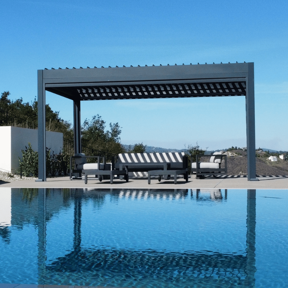 A dark gray Bon Pergola with a motorized louvered roof sits on a light gray platform, positioned next to a clear blue swimming pool. The louvers are partially open, allowing sunlight to filter through the deep gray slats, creating a bright and inviting outdoor space.