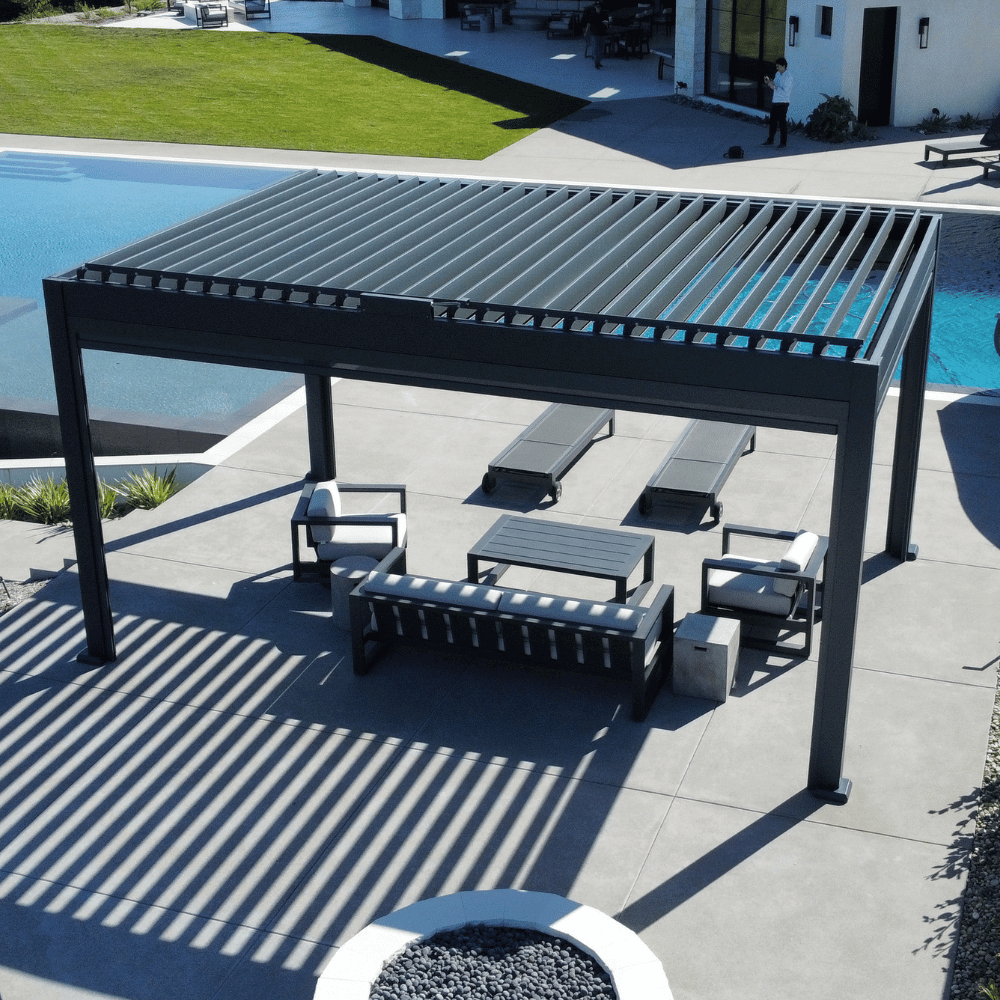 The dark gray Bon Pergola with a motorized louvered roof sits on a light gray platform, right next to a clear blue swimming pool. The overall atmosphere is luxurious, spacious, and resort-like. The dark gray pergola contrasts beautifully with the surrounding blue sky, lush greenery, and white architecture, creating a striking and inviting outdoor space.