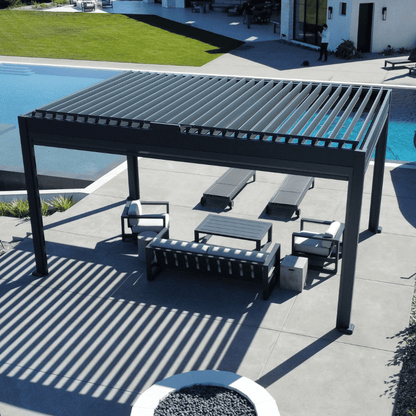 The dark gray Bon Pergola with a motorized louvered roof sits on a light gray platform, right next to a clear blue swimming pool. The overall atmosphere is luxurious, spacious, and resort-like. The dark gray pergola contrasts beautifully with the surrounding blue sky, lush greenery, and white architecture, creating a striking and inviting outdoor space.