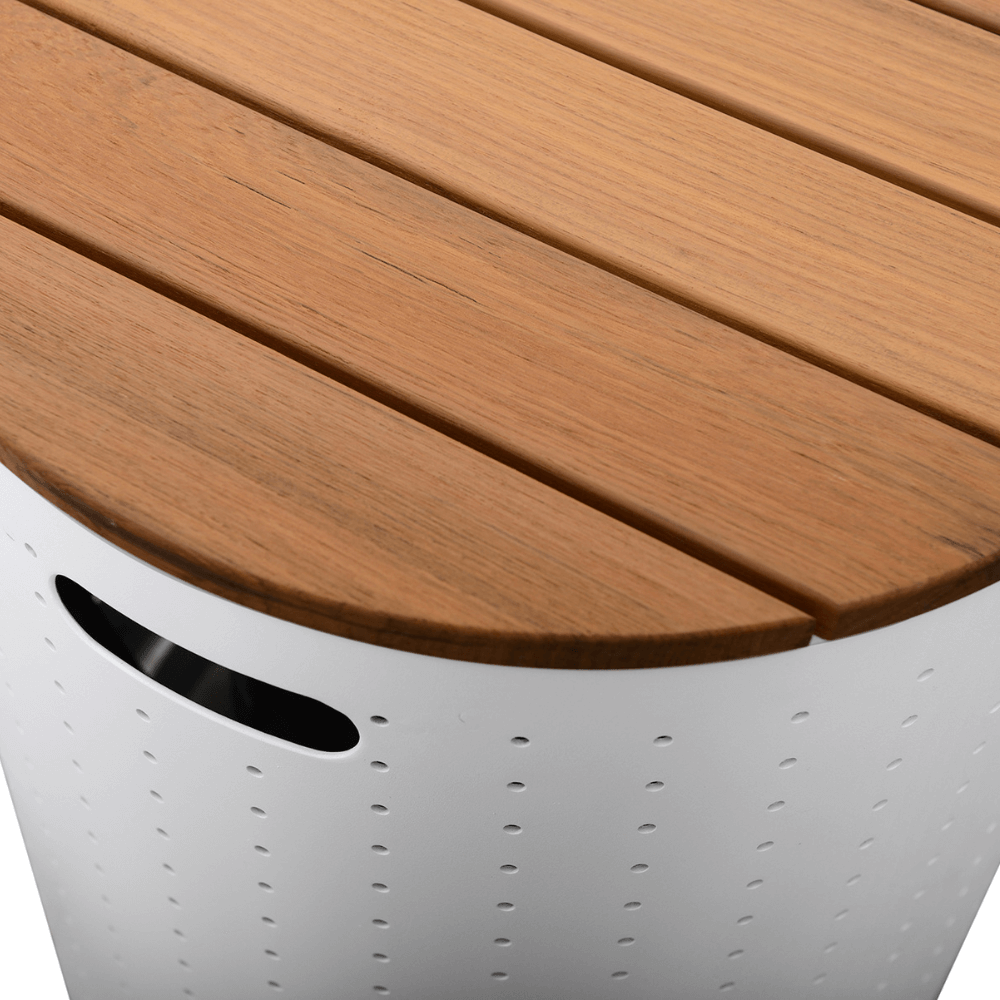 The storage box top is made of parallel light brown faux wood slats with subtle gaps between them, mimicking a wood grain texture. In the top-right corner of the image, a section of the faux wood tabletop is visible, clearly showing the texture and seams. The side of the storage box features white metal with small perforations, and a partial view of a rectangular handle opening is visible.