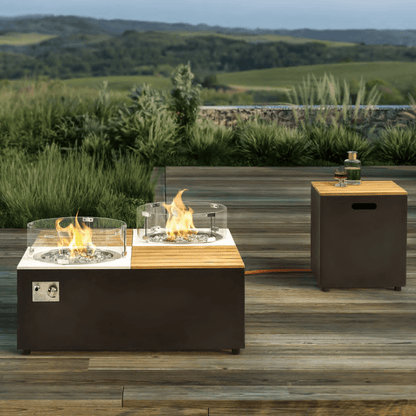 On the left is a square gas fire pit coffee table with a dark gray base and a light-colored faux wood slat tabletop. A round gas burner with flames burning is inset at each diagonal, surrounded by decorative stones and enclosed by a transparent glass wind guard. The side of the coffee table features a control panel. An orange gas line connects to a square deep gray storage box on the right, which also has a light-colored faux wood slat top and handle openings on the sides.