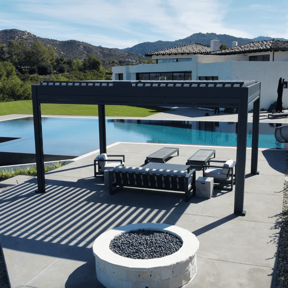 The dark gray Bon Pergola with a motorized louvered roof sits on a light gray platform, adjacent to a clear blue swimming pool. The louvers are partially open, casting distinct striped shadows below. The space is furnished with a modern outdoor lounge set, including a dark gray sofa, two armchairs, and a coffee table, complemented by light gray cushions. Additionally, two dark gray outdoor lounge chairs are placed on the platform, creating a relaxing and stylish outdoor retreat.