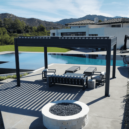 The dark gray Bon Pergola with a motorized louvered roof sits on a light gray platform, adjacent to a clear blue swimming pool. The louvers are partially open, casting distinct striped shadows below. The space is furnished with a modern outdoor lounge set, including a dark gray sofa, two armchairs, and a coffee table, complemented by light gray cushions. Additionally, two dark gray outdoor lounge chairs are placed on the platform, creating a relaxing and stylish outdoor retreat.