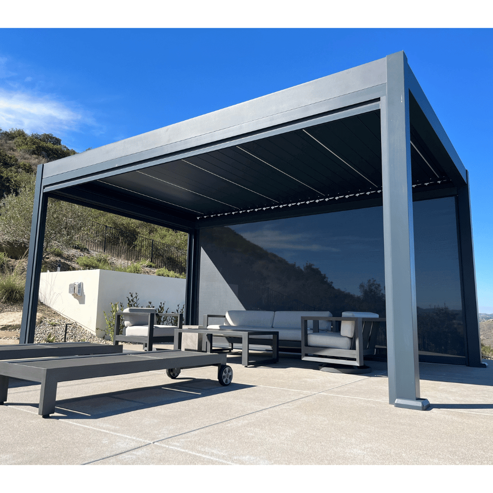 A dark gray Bon Pergola with a motorized louvered roof is positioned on a hillside or elevated area, offering views of the surrounding greenery and landscape. The sky is clear, displaying a vibrant blue color. The louvers' slat structure is visible, and the pergola is built with sturdy dark gray beams and posts. At least two sides of the pergola are fitted with dark shade screens, providing soft shading and enhancing privacy.