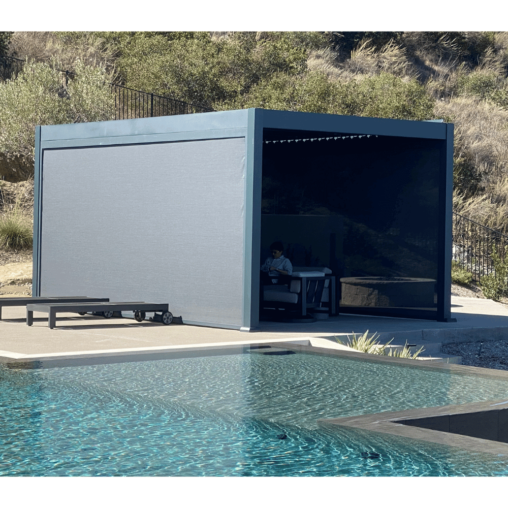 The dark gray Bon Pergola features closed louvers, creating a solid roof that provides full shade. Its sturdy frame is made of thick dark gray beams and posts, with windproof shade screens installed on both sides. Someone is seated inside, enjoying the sheltered space. The pergola sits on a platform with light gray flooring, positioned near the edge of a sleek black water feature or a low-profile pool.