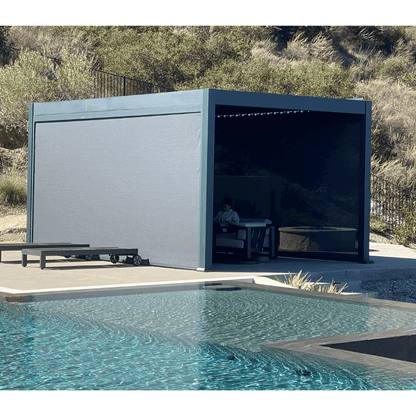 The dark gray Bon Pergola features closed louvers, creating a solid roof that provides full shade. Its sturdy frame is made of thick dark gray beams and posts, with windproof shade screens installed on both sides. Someone is seated inside, enjoying the sheltered space. The pergola sits on a platform with light gray flooring, positioned near the edge of a sleek black water feature or a low-profile pool.