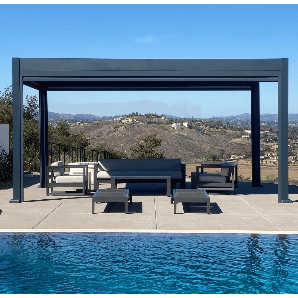 The dark gray Bon Pergola features a closed motorized louvered roof, creating a solid canopy for full shade. Its structure is made of thick dark gray beams and posts with a clean, modern design. Beneath the pergola is a stylish outdoor lounge set, including a sofa, two armchairs, and two small side tables. The cushions are light gray, adding a soft contrast to the sleek structure.