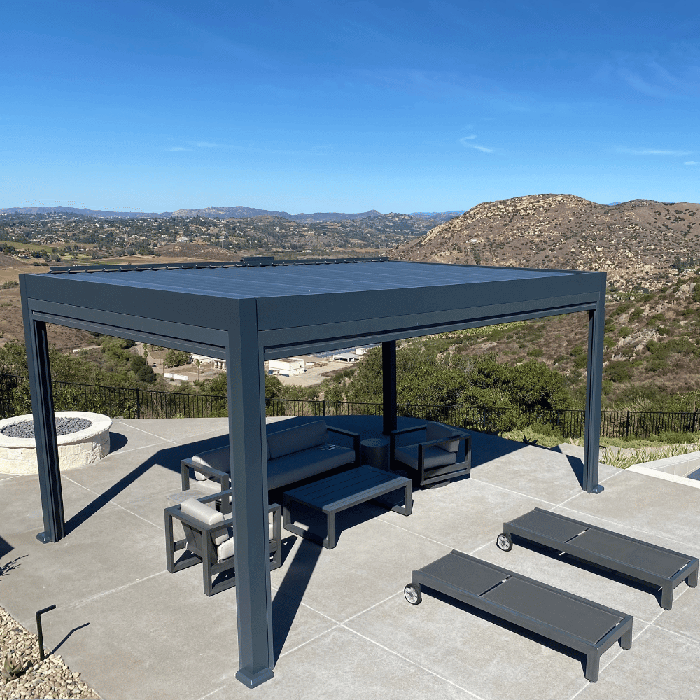 The black Bon Pergola features closed louvers, creating a solid black roof overhead. Below, a modern outdoor lounge set is arranged, along with two dark gray outdoor lounge chairs with wheels for easy movement. Positioned in a high location, the pergola offers stunning views, creating a relaxing outdoor space perfect for unwinding and enjoying the breathtaking scenery.
