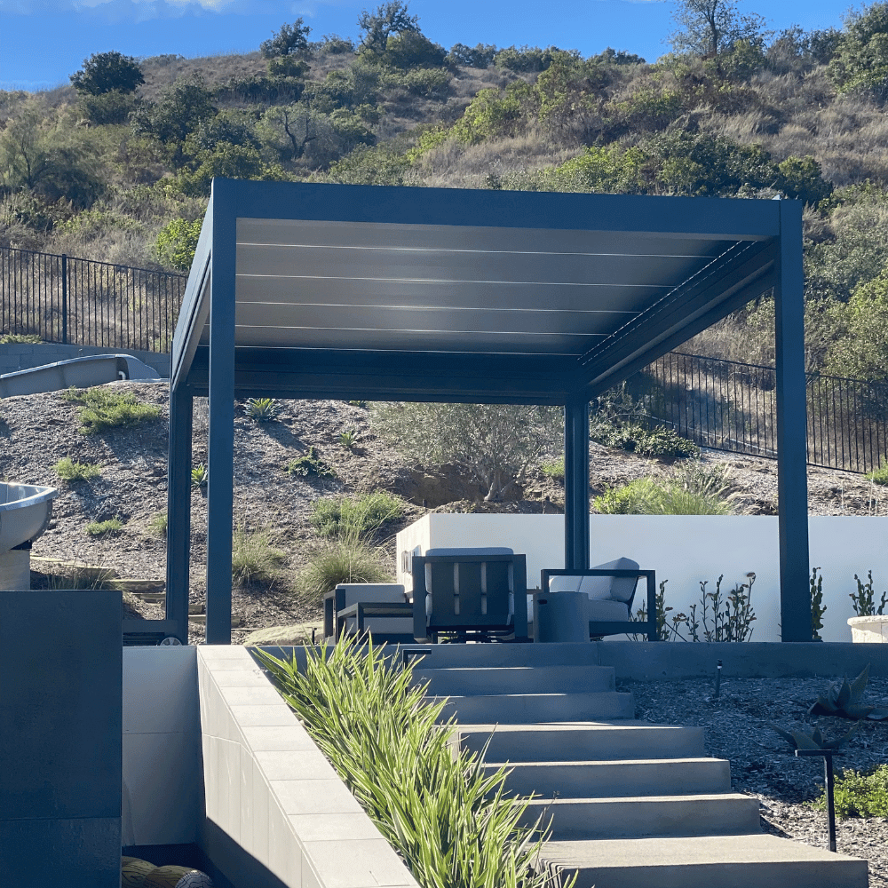 The dark gray Bon Pergola with a motorized louvered roof is situated atop a hill or elevated area, offering views of the surrounding greenery and terrain. The pergola is placed on a platform with light gray flooring, connected to the lower area by several steps made of gray stone. Surrounding the pergola is lush, green vegetation and rolling hills, enhancing the peaceful, natural atmosphere.