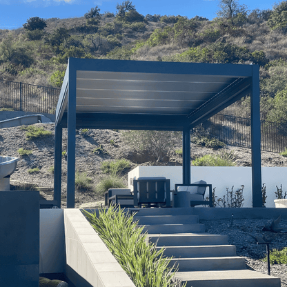 The dark gray Bon Pergola with a motorized louvered roof is situated atop a hill or elevated area, offering views of the surrounding greenery and terrain. The pergola is placed on a platform with light gray flooring, connected to the lower area by several steps made of gray stone. Surrounding the pergola is lush, green vegetation and rolling hills, enhancing the peaceful, natural atmosphere.
