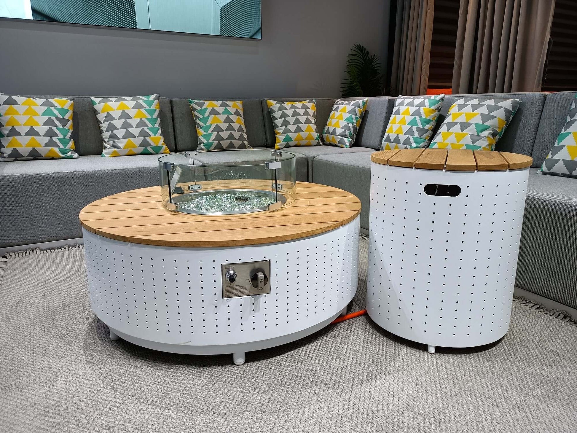 The DRIGANI outdoor gas fire pit coffee table and matching side table feature a white metal base with a surface covered in circular holes. The tabletop is made of light wood-grain metal panels, with a round gas fire pit embedded in the center, surrounded by a transparent circular glass wind guard. On the side of the coffee table, there's a metal control panel with a knob and ignition system, and an orange gas hose extends from the bottom of the table.