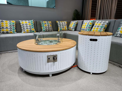 The DRIGANI outdoor gas fire pit coffee table and matching side table feature a white metal base with a surface covered in circular holes. The tabletop is made of light wood-grain metal panels, with a round gas fire pit embedded in the center, surrounded by a transparent circular glass wind guard. On the side of the coffee table, there's a metal control panel with a knob and ignition system, and an orange gas hose extends from the bottom of the table.