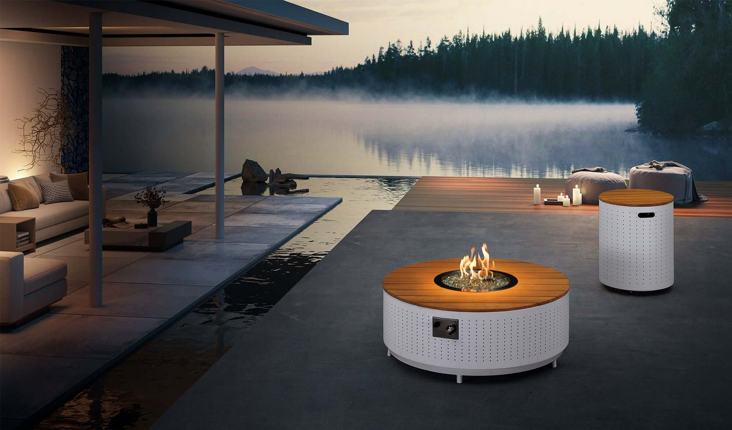 The round Cypress Collection Fire Pit coffee table features a white perforated metal base and a light-colored faux wood slat tabletop. At the center, a circular gas fire pit burns bright flames, surrounded by white decorative stones. A metal control panel is located on the side. To the rear right, a cylindrical propane tank storage box matches the table’s base, with a top made of light wood slats and a rectangular handle opening on the side.