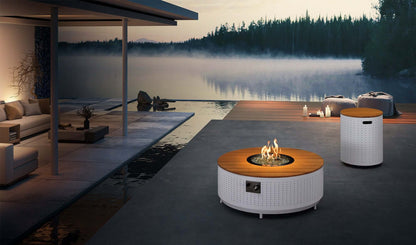 The round Cypress Collection Fire Pit coffee table features a white perforated metal base and a light-colored faux wood slat tabletop. At the center, a circular gas fire pit burns bright flames, surrounded by white decorative stones. A metal control panel is located on the side. To the rear right, a cylindrical propane tank storage box matches the table’s base, with a top made of light wood slats and a rectangular handle opening on the side.