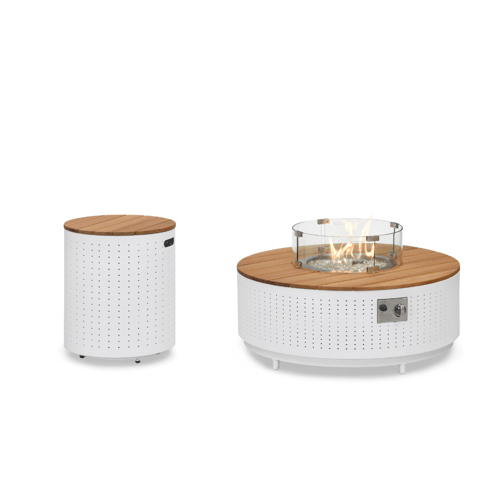 The Cypress Collection Fire Pit and matching storage box feature a white perforated metal base with a light faux wood slat tabletop. The coffee table has a circular gas fire pit in the center, surrounded by transparent decorative stones and enclosed by a clear glass wind guard. A metal control panel is on the side. To the left, a cylindrical storage box matches the table's base, with a light wood slat top and a rectangular handle opening on the side.