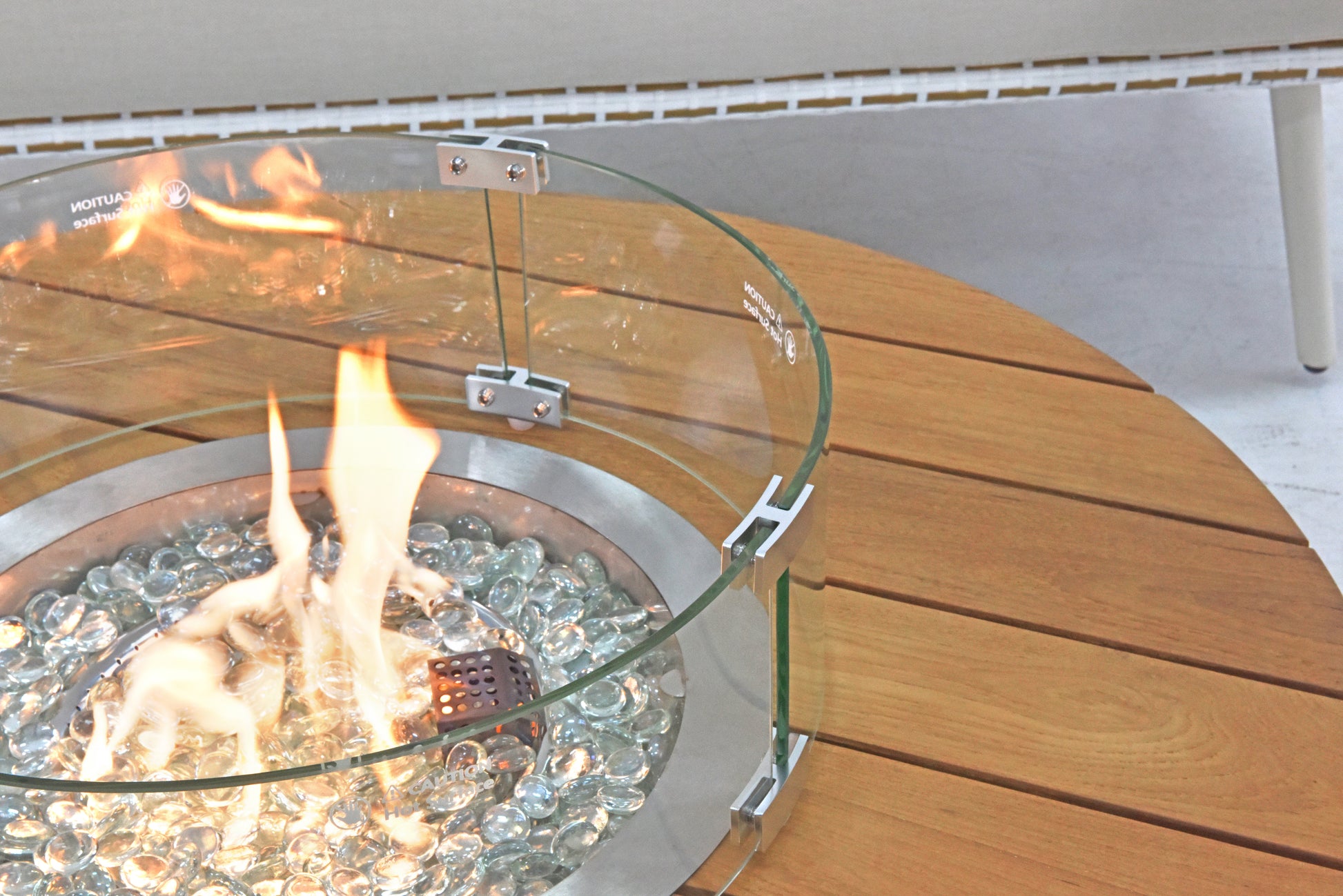 The outdoor gas fire pit is positioned on a wooden tabletop, surrounded by a curved transparent glass wind guard that combines both functionality and aesthetic appeal.