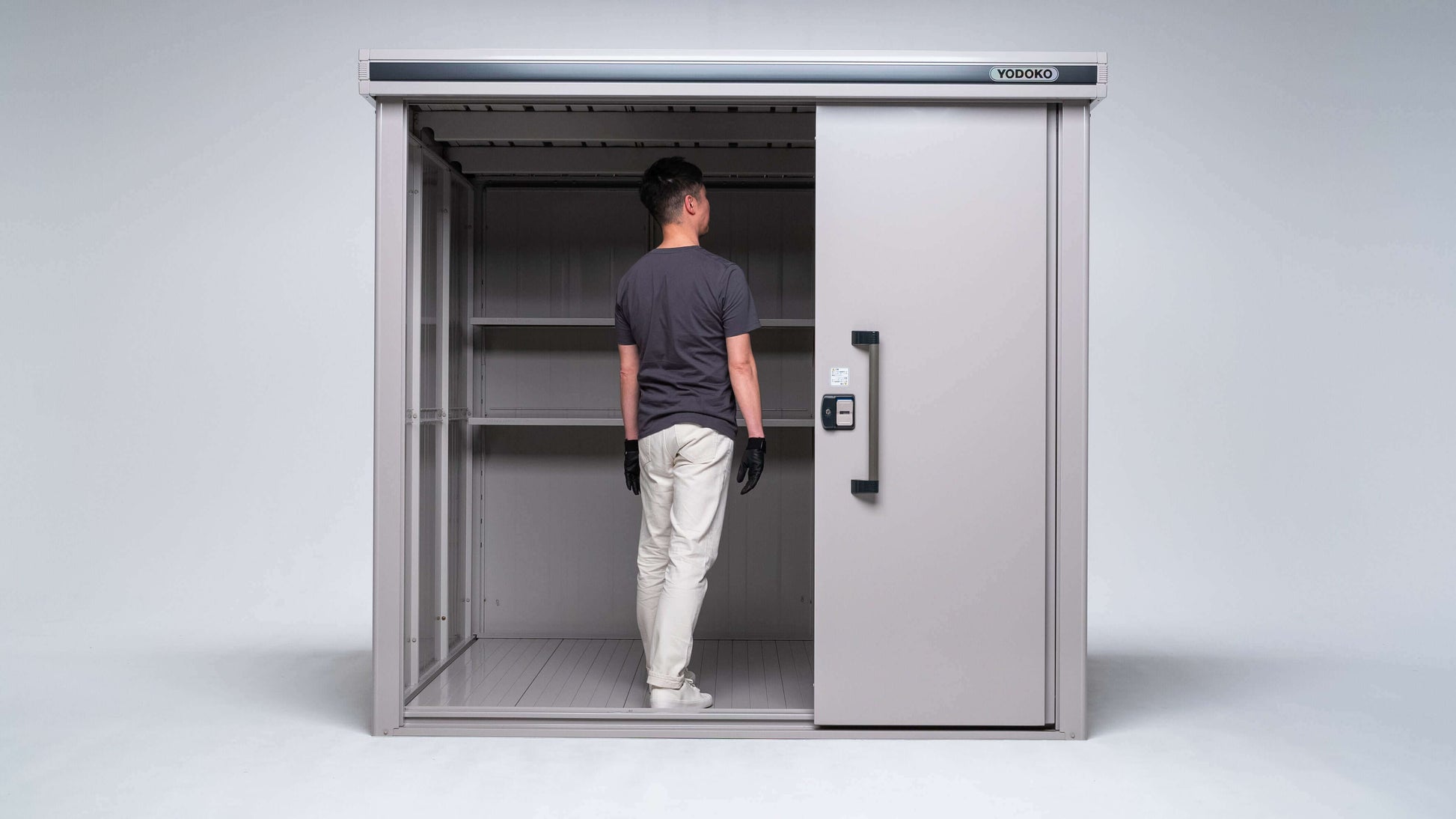 A man stands inside a large, open YODOKO Premium Outdoor Large Shed LMDS by BON PERGOLA, showcasing Japanese craftsmanship. He faces sideways in a dark shirt, light pants, and gloves. The metallic shed features a sliding door and durable coating on its empty shelves and smooth floor.