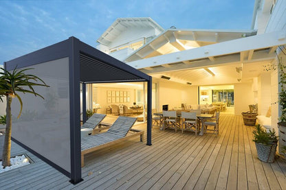The black motorized louvered pergola has its louvers fully closed, creating a solid roof. Light gray shade screens on the sides offer both privacy and sun protection. Beneath the pergola are two wooden lounge chairs and a small side table—perfect for relaxing day or night. Soft indoor lighting contrasts with the evening sky, creating a peaceful, serene vibe.