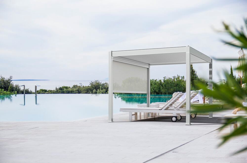 A white Bon Pergola sits poolside in a luxury residential area, featuring a sleek fixed canopy that provides continuous shade. Its slim white posts and beams create a clean, modern silhouette. A manually retractable white shade on one side is partially lowered for added sun protection and privacy. The overall vibe is serene, elegant, and resort-like.