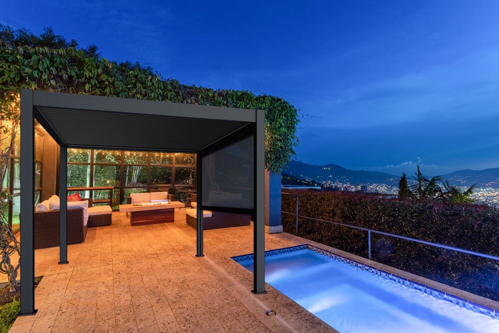 The outdoor lounge area features a 10'x13' deep gray Bon Pergola with motorized louvers. The structure is made of deep gray posts and beams, with warm yellow LED lights along the edges of the beams, casting a soft glow over the area beneath. Next to the pergola is a terrace with light brown floor tiles, and on one side, a rectangular swimming pool whose surface reflects a soft blue hue at night, enhancing the tranquil atmosphere.