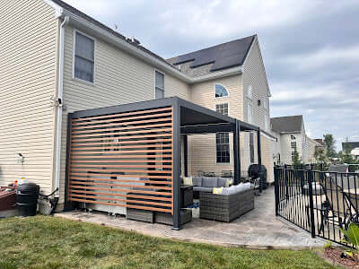 The pergola is positioned right up against the beige exterior wall of the house, with windows and a black roof creating a modern, clean visual contrast, making for a cozy outdoor relaxation space.