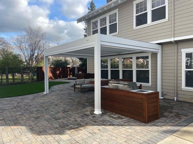 The white Bon Pergola features partially open louvers, with a sturdy frame of thick white beams and posts. Below, a patio paved in brown and gray tiles hosts a light gray outdoor sofa with a wooden frame, paired with a dark brown wooden outdoor kitchen or bar counter. The counter is equipped with a grill and sink, perfect for outdoor cooking and entertaining.