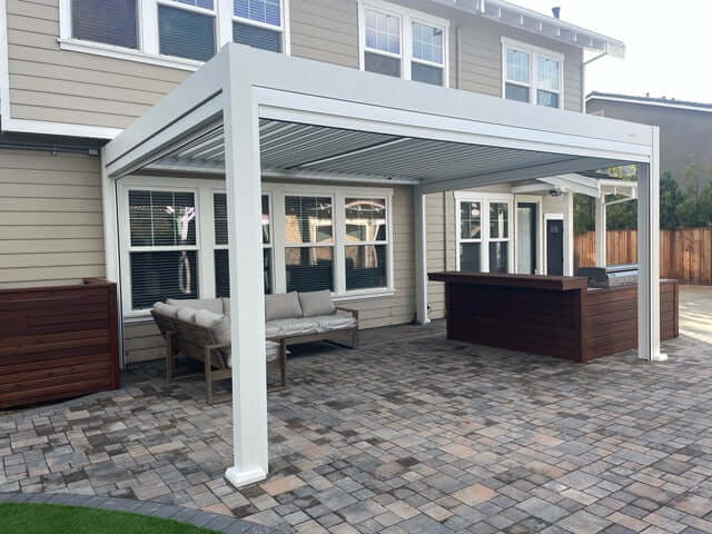 The pergola features thick white posts and beams in a clean, modern design, positioned right against the back wall of the house, which has multiple windows. Below is a patio paved with a mix of brown and gray tiles. A light gray outdoor corner sofa with a wooden frame sits on the patio, while one side features a dark brown wooden bar or outdoor kitchen counter.