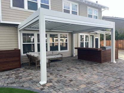The pergola features thick white posts and beams in a clean, modern design, positioned right against the back wall of the house, which has multiple windows. Below is a patio paved with a mix of brown and gray tiles. A light gray outdoor corner sofa with a wooden frame sits on the patio, while one side features a dark brown wooden bar or outdoor kitchen counter.