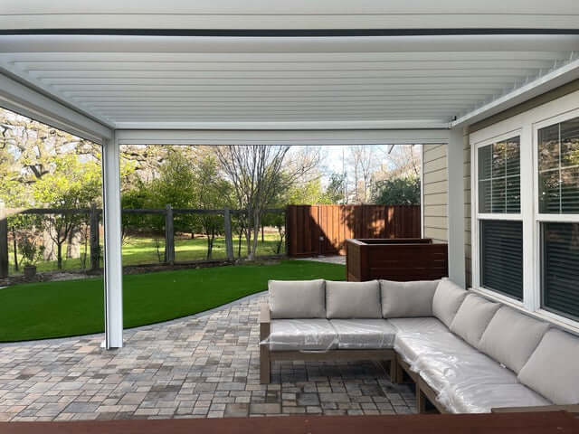 The white Bon Pergola features closed louvers, forming a solid roof that offers full shade. Its structure includes thick white columns and beams with a clean, modern design. The pergola is attached to the back wall of the house, which has one window framed with white trim. The home’s exterior is clad in light brown siding, creating a warm, inviting contrast.