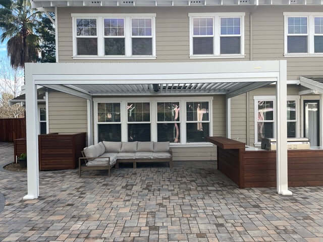A sleek white Bon Pergola with partially open louvered roof casts slim, striped shadows below. Its sturdy columns and beams create a clean, modern look, attached directly to the back wall of the house lined with several windows. Beneath it lies a patio paved in a mix of brown and gray tiles. A light gray outdoor sectional with a wooden frame sits comfortably on the patio, flanked by dark brown wooden storage units on either side.