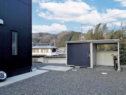 An outdoor scene features a small black BON PERGOLA building on the left, known for its durable coating, and a YODOKO Premium Outdoor Large Shed LMDS on the right with heat-insulated technology housing a bicycle. Gravel covers the ground, with trees and buildings under a partly cloudy sky.