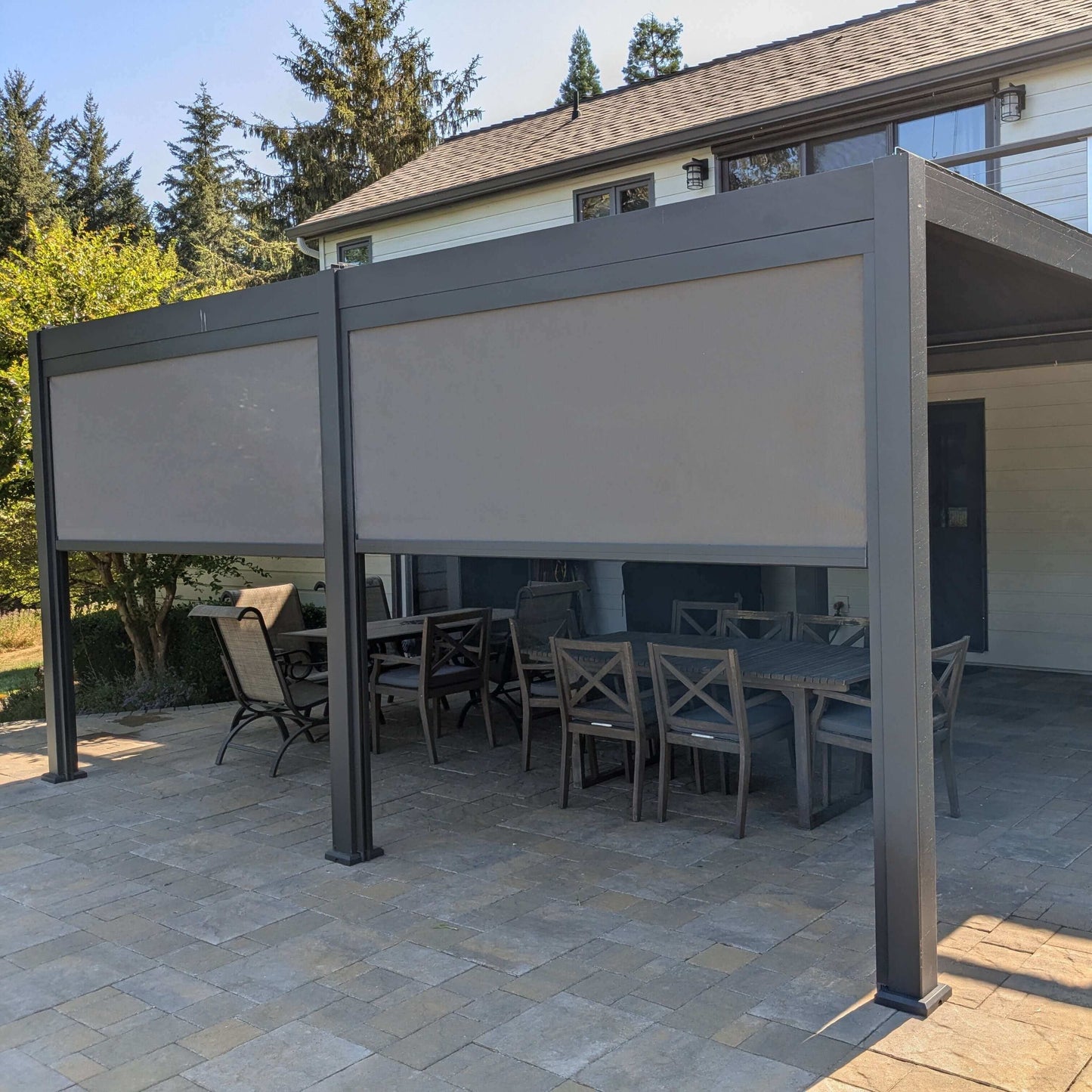 Installed on a backyard patio, the deep gray Villa Pergola features a closed louvered roof, forming a solid overhead cover. Its structure is made of sturdy deep gray posts and beams with a modern look. Light gray motorized blinds are mounted on the sides, some partially lowered to provide shade and privacy. The blinds appear semi-translucent, effectively blocking direct sunlight while still letting in soft, filtered light.