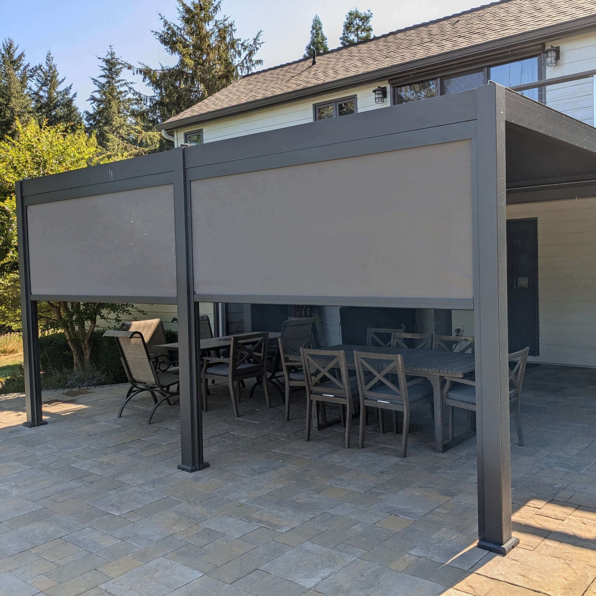Installed on a backyard patio, the deep gray Villa Pergola features a closed louvered roof, forming a solid overhead cover. Its structure is made of sturdy deep gray posts and beams with a modern look. Light gray motorized blinds are mounted on the sides, some partially lowered to provide shade and privacy. The blinds appear semi-translucent, effectively blocking direct sunlight while still letting in soft, filtered light.