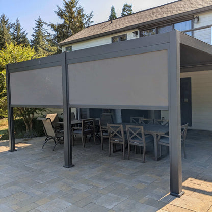 Installed on a backyard patio, the deep gray Villa Pergola features a closed louvered roof, forming a solid overhead cover. Its structure is made of sturdy deep gray posts and beams with a modern look. Light gray motorized blinds are mounted on the sides, some partially lowered to provide shade and privacy. The blinds appear semi-translucent, effectively blocking direct sunlight while still letting in soft, filtered light.