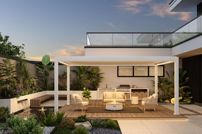 This is a white Louvre Roof pergola with the louvers partially open, featuring a warm LED light strip along the edge of the top. Beneath the pergola is a cozy outdoor lounge area with wooden flooring, furnished with a beige two-person sofa, two beige woven armchairs, and a round coffee table with a gas fire pit in the center. Behind the seating area is a modern outdoor kitchen, complete with light-colored countertops, a stainless steel sink, a grill, and storage cabinets. The entire space combines shading, 