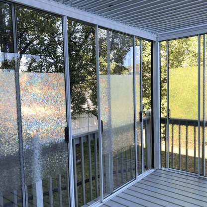 The enclosed outdoor terrace or balcony features a white louvered pergola with partially open slats, revealing the louver structure. The pergola's support structure is also white. The sides are equipped with multiple white-framed sliding glass doors, separating the terrace from the outdoors and creating a semi-indoor space. Some of the glass doors are decorated with rainbow-colored film.