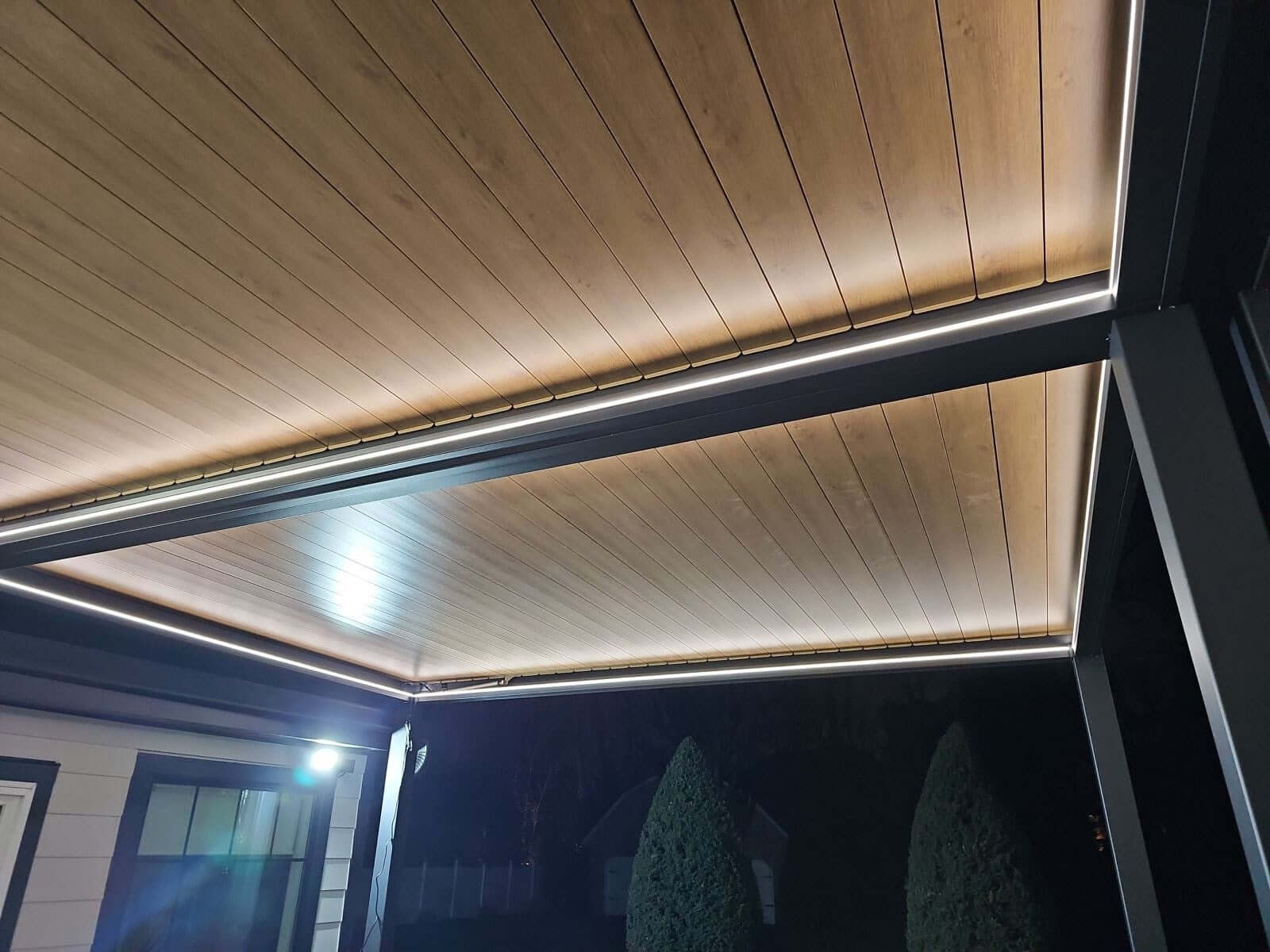 This is a close-up of the pergola roof. The roof is made up of several parallel slats with a wood-like finish, supported by a simple black frame. A white LED strip runs along the inner edge of the roof, casting a bright, even light that illuminates the space below and clearly highlights the pergola’s shape, creating a striking contrast with the dark frame.