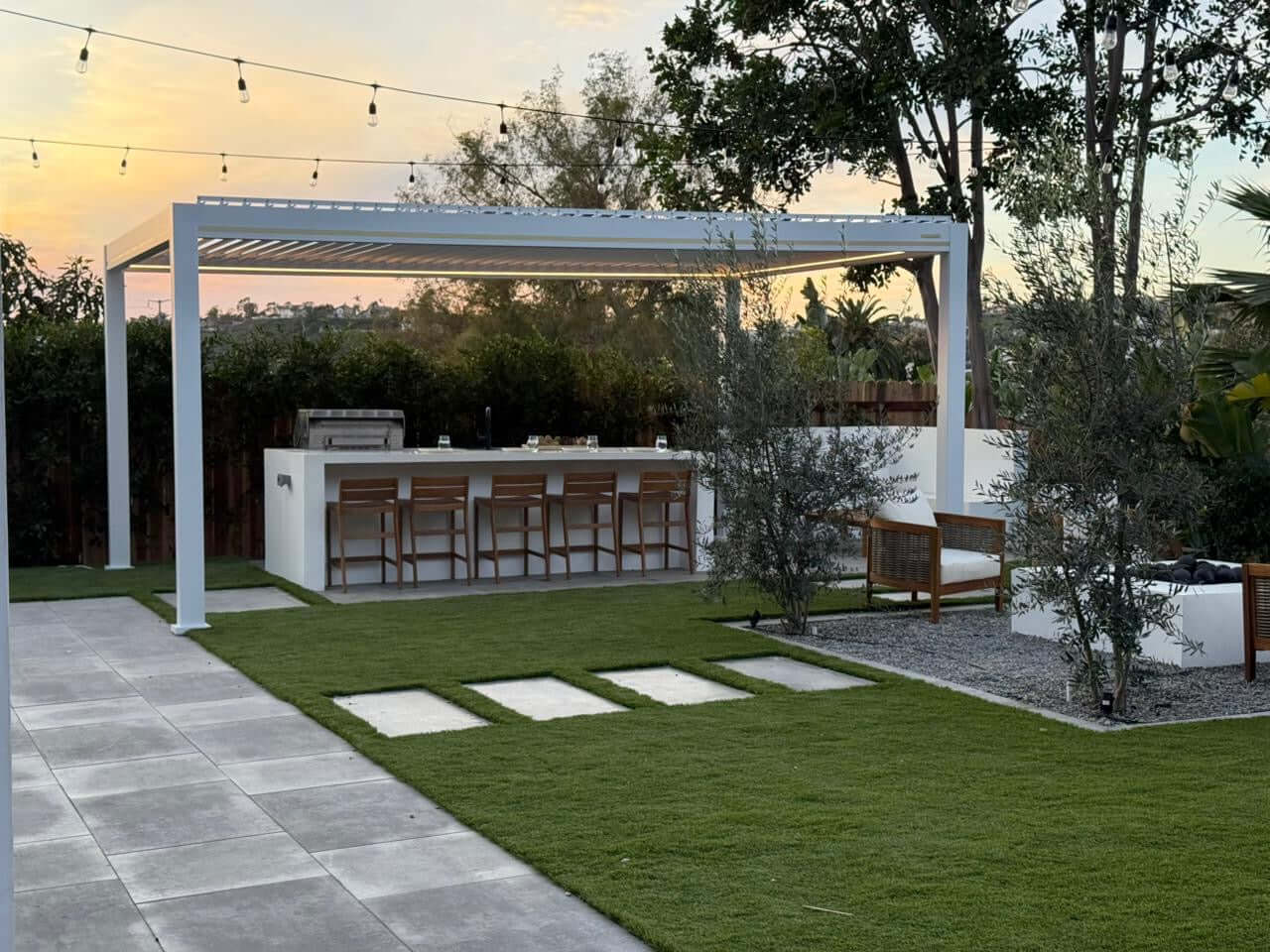 A white motorized louvered pergola with the louvers partially open. Below it is an outdoor bar area with a light-colored countertop and five wooden bar stools. Behind the bar is an outdoor grill, making this space perfect for both relaxing and cooking outdoors.