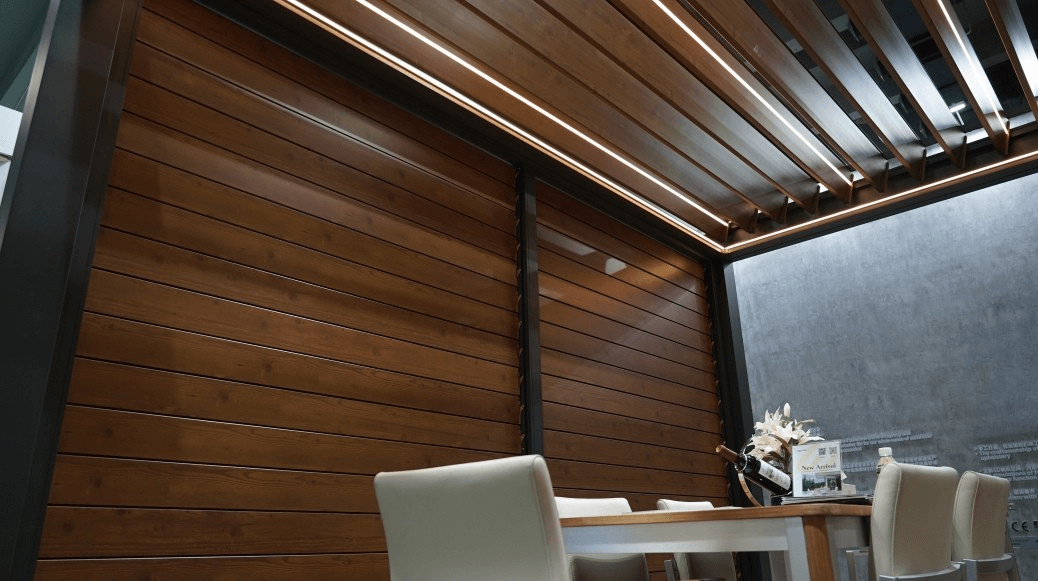 The pergola features a dark frame with a wood grain louver roof. The wooden grain louver can be opened to reveal glimpses of the sky. Below the pergola is a dining area, with a wooden table and light-colored chairs. The wood grain slat walls offer partial enclosure, adding a sense of privacy.