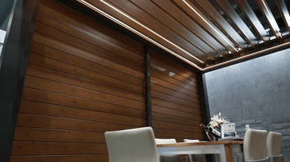 The pergola features a dark frame with a wood grain louver roof. The wooden grain louver can be opened to reveal glimpses of the sky. Below the pergola is a dining area, with a wooden table and light-colored chairs. The wood grain slat walls offer partial enclosure, adding a sense of privacy.