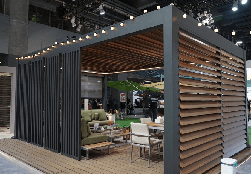 An outdoor pavilion features a sleek design similar to the Custom Weatherproof Pergola, with string lights above. Green seating complements white chairs around a low table amid stylish decor, while green umbrellas offer shade to relaxed visitors in the background.