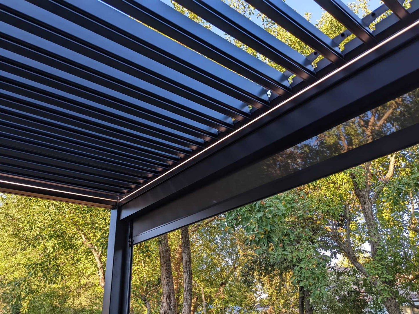 The dark motorized louvered pergola features partially open louvers that cast slender linear shadows. Its structure is made of dark pillars and beams, with warm yellow LED strips subtly glowing along the edges. A dark shade screen on one side is partially lowered for added sun protection.